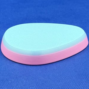 May include: A light blue and pink oval-shaped ceramic coaster. The coaster has a smooth, slightly textured surface.