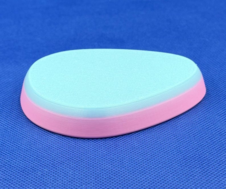 May include: A light blue and pink oval-shaped ceramic coaster. The coaster has a smooth, slightly textured surface.