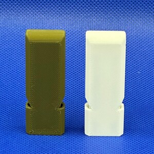 Op de afbeelding: Twee rechthoekige 3D-geprinte fluitjes, &eacute;&eacute;n in olijfgroen en &eacute;&eacute;n in off-white, met een platte bovenkant, een taps toelopend middengedeelte en een bredere basis met een zichtbare opening voor de klankkast onderaan.