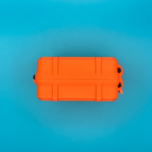 May include: A rectangular, bright orange protective storage case featuring a reinforced, ribbed exterior design. The case is secured with small black screws visible on the side hinges, providing a sturdy and compact enclosure for items.