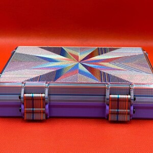 May include: A rectangular storage case featuring a vibrant, multi-colored geometric starburst pattern on the lid. The sides and two front latches display a layered, striped texture in shades of blue, purple, and orange.