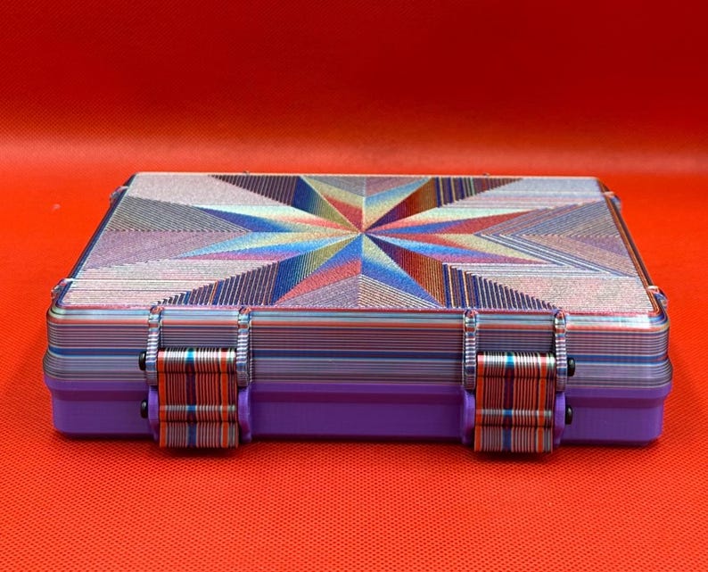 May include: A rectangular storage case featuring a vibrant, multi-colored geometric starburst pattern on the lid. The sides and two front latches display a layered, striped texture in shades of blue, purple, and orange.