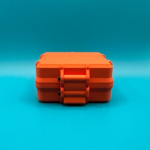 May include: A bright orange rectangular storage case with a textured matte finish. The case features a secure latch mechanism on the front with two small black pins visible at the hinge points, providing a compact and durable design.