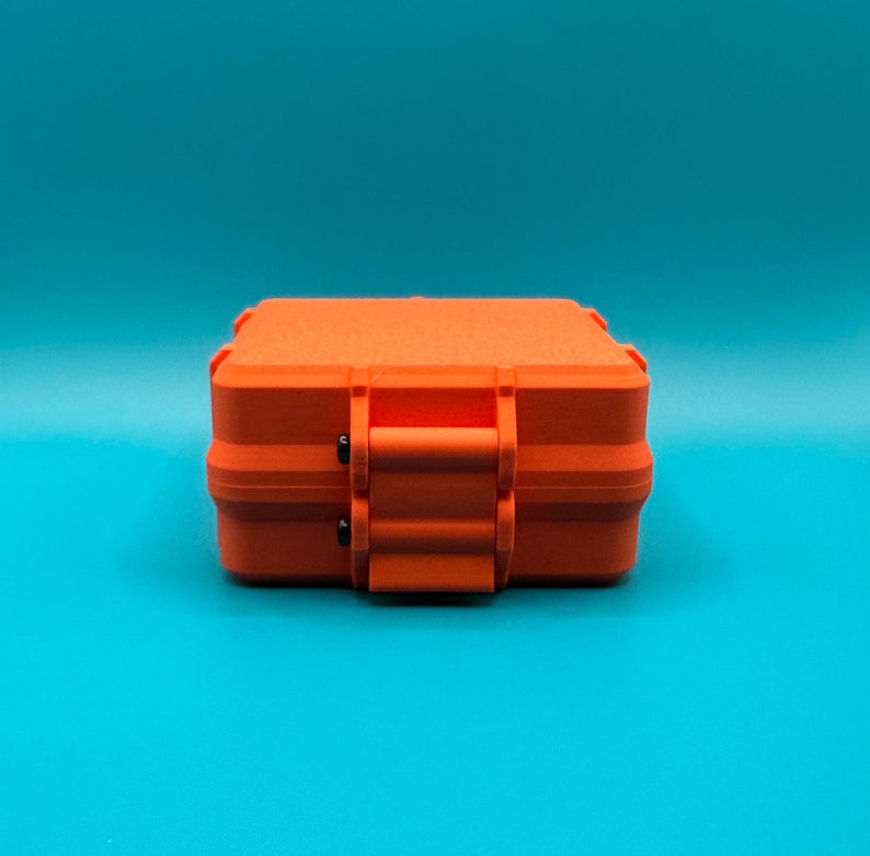 May include: A bright orange rectangular storage case with a textured matte finish. The case features a secure latch mechanism on the front with two small black pins visible at the hinge points, providing a compact and durable design.