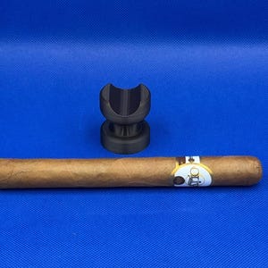 May include: A black cigar rest with a brown cigar resting on top. The cigar has a gold band with the word "Olds" printed on it.