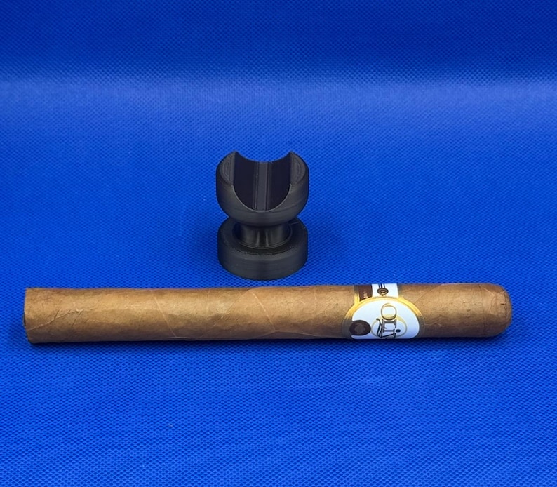 May include: A black cigar rest with a brown cigar resting on top. The cigar has a gold band with the word "Olds" printed on it.