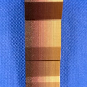 May include: A close-up of a copper-colored metal sheet with a brushed finish. The sheet is bent at a 90-degree angle, creating a sharp edge. The surface has a subtle gradient of color, transitioning from a darker copper hue to a lighter gold tone.