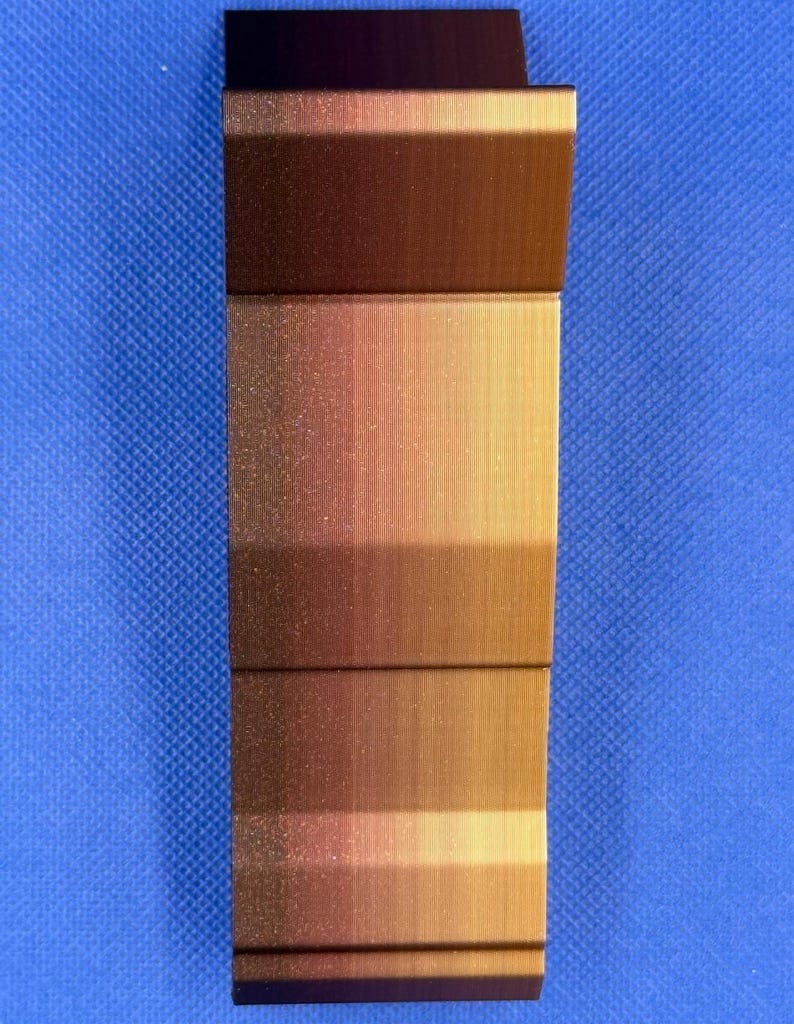 May include: A close-up of a copper-colored metal sheet with a brushed finish. The sheet is bent at a 90-degree angle, creating a sharp edge. The surface has a subtle gradient of color, transitioning from a darker copper hue to a lighter gold tone.