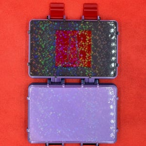 May include: Open, purple, rectangular case with a holographic star pattern on the inside. The top section has a red square with a star pattern. The case is open, revealing the interior. The background is a solid red.