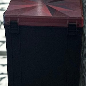 May include: A black box with a red and black geometric design on the lid. The lid has a sunburst pattern in shades of red. The box has a rectangular shape and a hinged lid with a clasp.