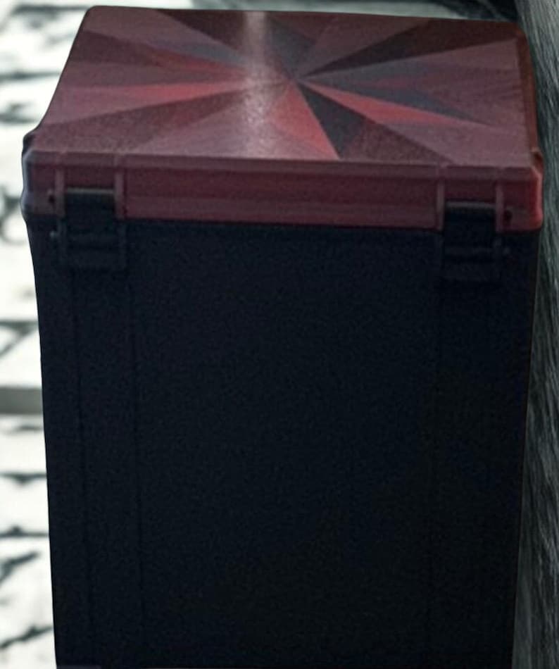 May include: A black box with a red and black geometric design on the lid. The lid has a sunburst pattern in shades of red. The box has a rectangular shape and a hinged lid with a clasp.