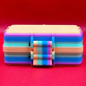 Puede incluir: Una caja de almacenamiento rectangular con un degradado de rayas horizontales en tonos morado, azul, turquesa y melocot&oacute;n. La caja tiene una tapa texturizada de color claro y un cierre central que coincide con el patr&oacute;n de rayas.