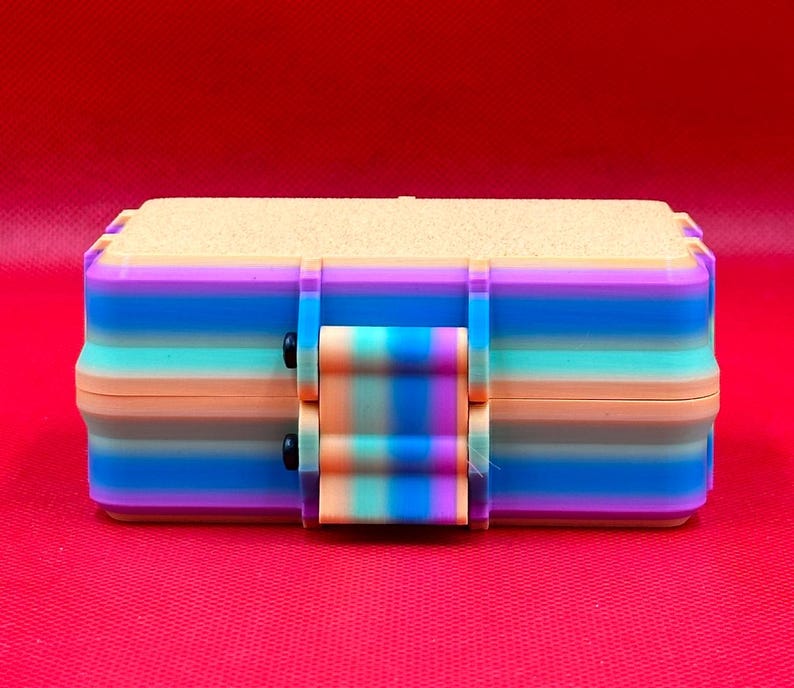 Puede incluir: Una caja de almacenamiento rectangular con un degradado de rayas horizontales en tonos morado, azul, turquesa y melocot&oacute;n. La caja tiene una tapa texturizada de color claro y un cierre central que coincide con el patr&oacute;n de rayas.