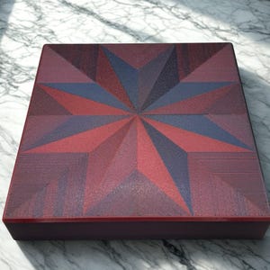 May include: A square wooden box with a geometric starburst design. The starburst features shades of red and blue. The box has a dark red border and is sitting on a marble surface.