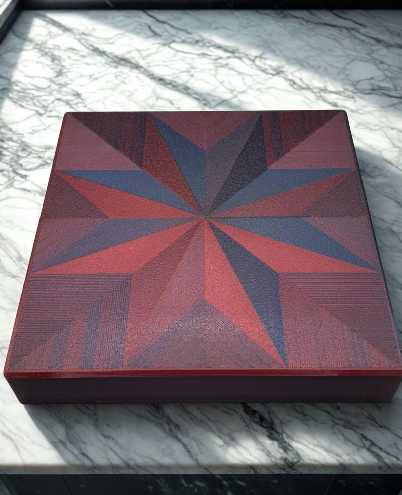 May include: A square wooden box with a geometric starburst design. The starburst features shades of red and blue. The box has a dark red border and is sitting on a marble surface.