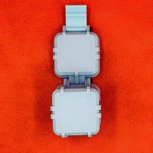 May include: A light blue plastic folding device featuring two square, flat-topped compartments connected by a central hinge mechanism, with a vertical tab extending from the top section, presented against a solid, vibrant orange background.