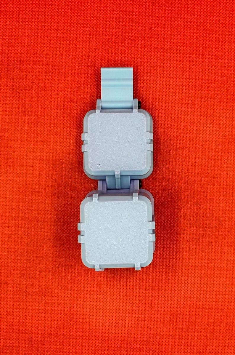 May include: A light blue plastic folding device featuring two square, flat-topped compartments connected by a central hinge mechanism, with a vertical tab extending from the top section, presented against a solid, vibrant orange background.