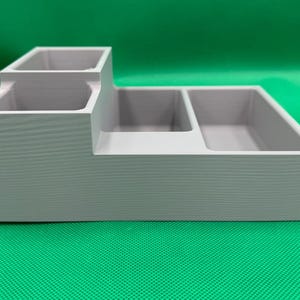 Op de afbeelding: Een grijze plastic organizer met drie vakken. De organizer heeft een verhoogd gedeelte met twee kleinere vakken en een groter vak aan de voorkant.