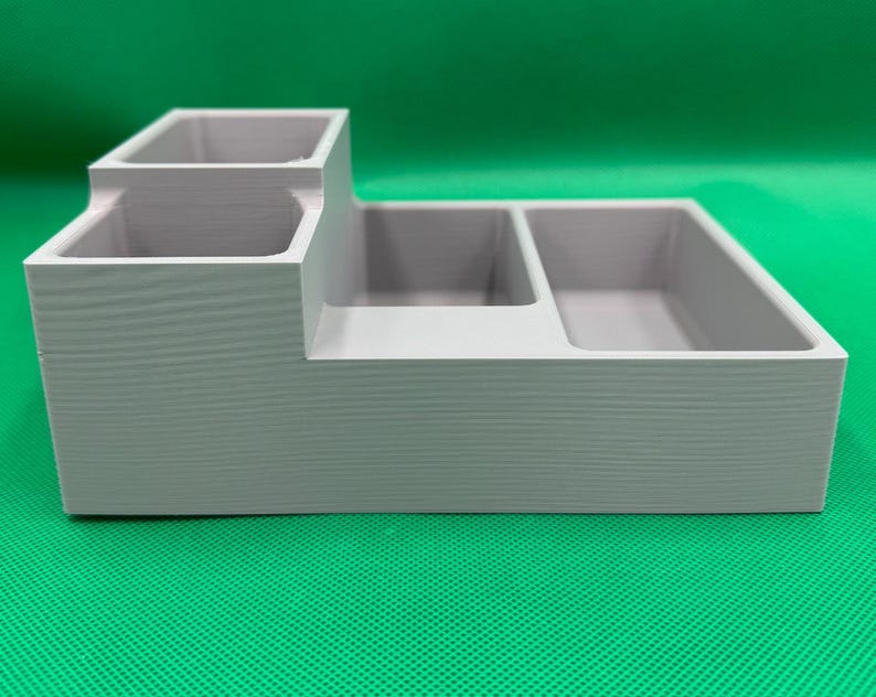 Op de afbeelding: Een grijze plastic organizer met drie vakken. De organizer heeft een verhoogd gedeelte met twee kleinere vakken en een groter vak aan de voorkant.