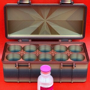 Puede incluir: Una caja de almacenamiento rectangular impresa en 3D con tapa abatible y diez compartimentos circulares, con un degradado de marr&oacute;n, negro y azul. Delante hay un vial de vidrio transparente con una tapa de color rosa.