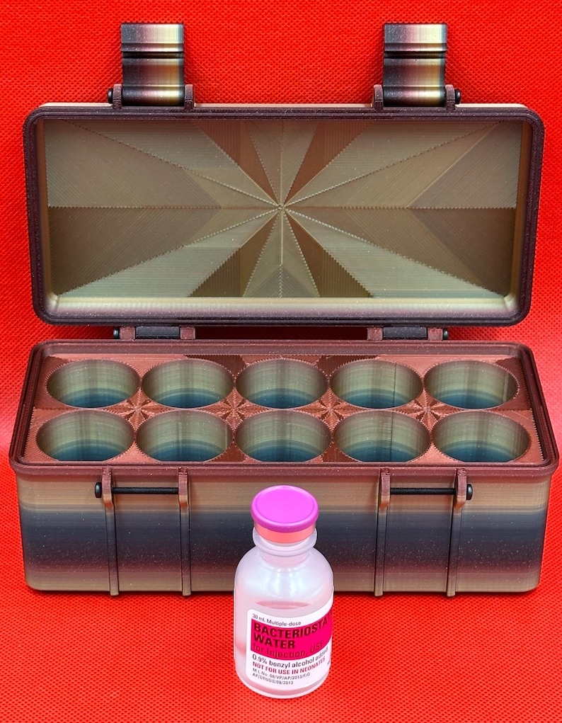 Puede incluir: Una caja de almacenamiento rectangular impresa en 3D con tapa abatible y diez compartimentos circulares, con un degradado de marr&oacute;n, negro y azul. Delante hay un vial de vidrio transparente con una tapa de color rosa.
