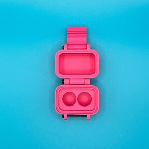 May include: A bright pink, rectangular, hinged plastic case shown in an open position. The top section is a hollow rectangular compartment, while the bottom section features two circular indentations. The case has a vertical orientation.