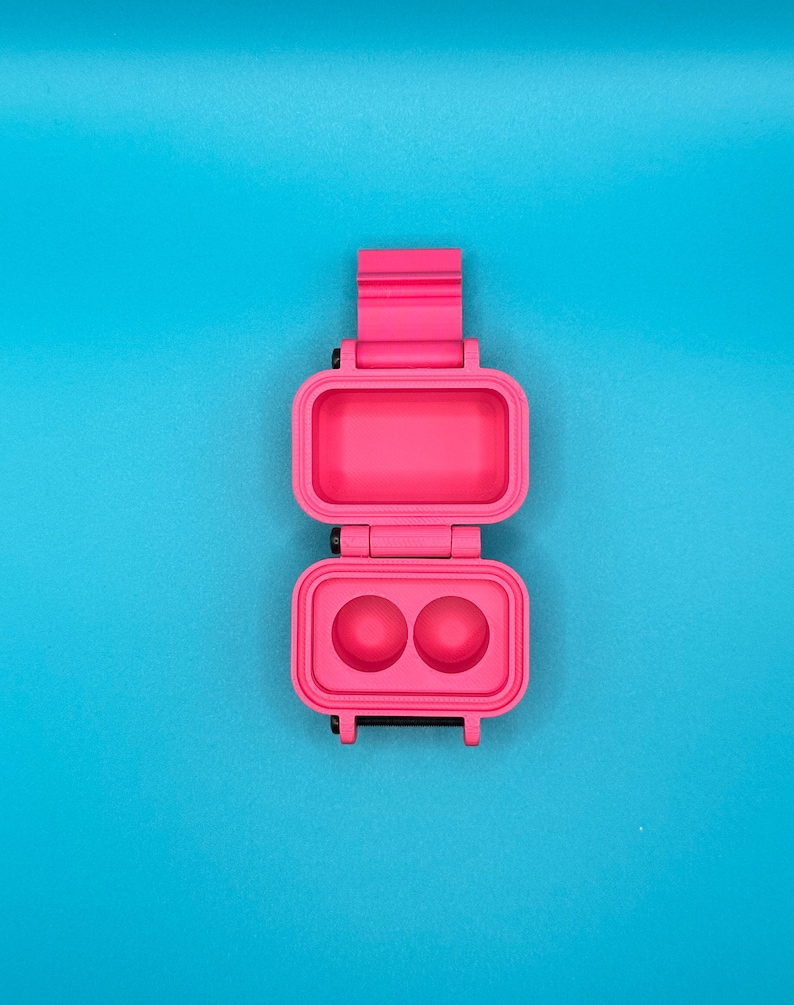 May include: A bright pink, rectangular, hinged plastic case shown in an open position. The top section is a hollow rectangular compartment, while the bottom section features two circular indentations. The case has a vertical orientation.