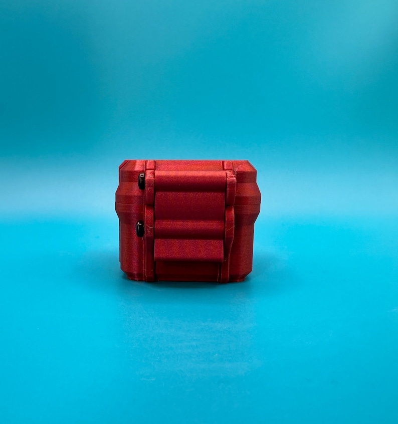 Puede incluir: Una peque&ntilde;a caja de almacenamiento rectangular de color rojo met&aacute;lico con un acabado texturizado. La caja presenta un dise&ntilde;o de tapa con bisagras y dos cierres circulares negros visibles en la parte frontal para asegurar el contenedor.