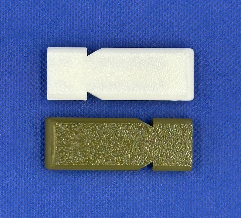 Op de afbeelding: Twee rechthoekige kunststof onderdelen met een getextureerd oppervlak en een ingekeepte zijkant. Het ene onderdeel is effen wit en het andere is olijfgroen. Beide items hebben een platte, langwerpige vorm met een duidelijke inkeping.