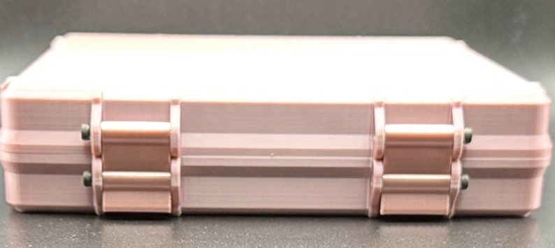 May include: A rectangular, light pink storage case featuring a smooth, solid-colored exterior. The case is constructed with two prominent hinges on the front side, each secured with small, dark-colored pins, providing a functional closure mechanism.