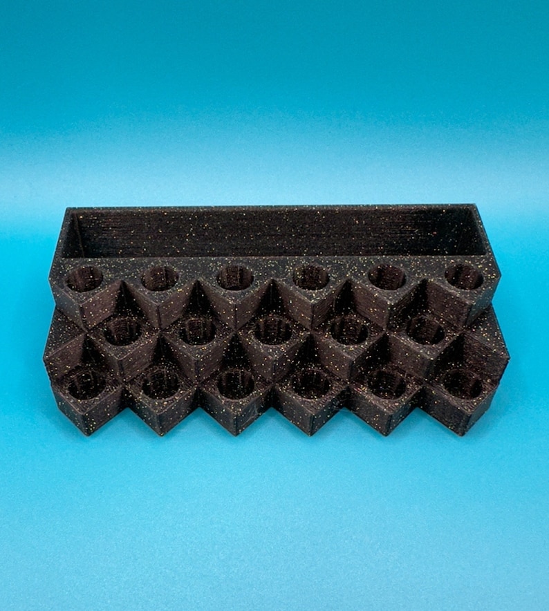 May include: A black, speckled, multi-tiered organizer stand featuring a rectangular storage compartment at the back and a series of twenty-one circular, recessed slots arranged in a grid pattern across the front stepped levels.
