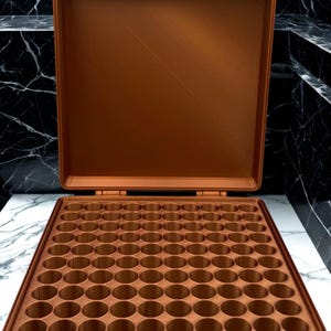 May include: A copper-colored, rectangular storage case with an open lid, featuring a grid of numerous circular compartments designed for organizing small items. The case has a sturdy hinge mechanism and a flat, smooth interior surface.