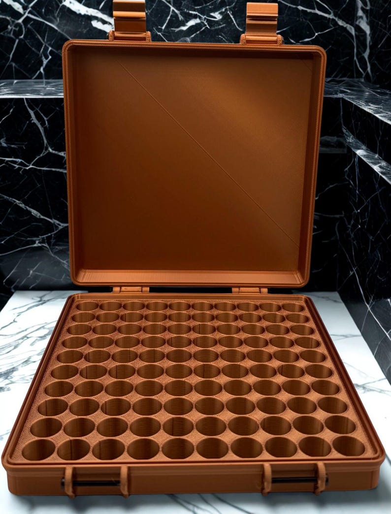 May include: A copper-colored, rectangular storage case with an open lid, featuring a grid of numerous circular compartments designed for organizing small items. The case has a sturdy hinge mechanism and a flat, smooth interior surface.
