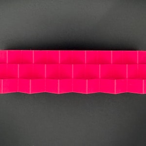 May include: A bright magenta display stand featuring a tiered, stepped design with multiple horizontal levels. The structure has a repeating geometric, zigzag pattern along the front edge, creating a series of small, uniform platforms.