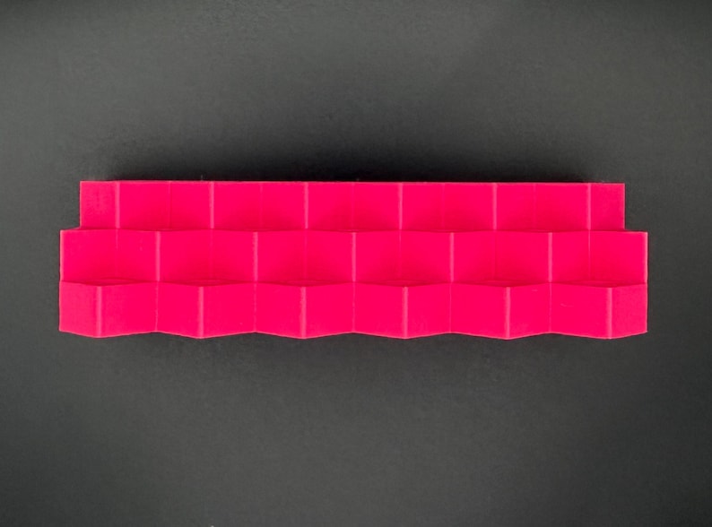 May include: A bright magenta display stand featuring a tiered, stepped design with multiple horizontal levels. The structure has a repeating geometric, zigzag pattern along the front edge, creating a series of small, uniform platforms.
