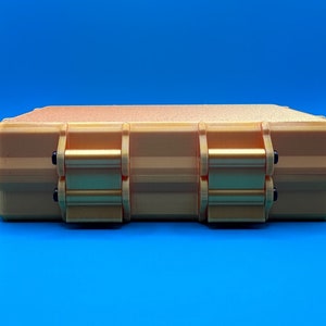 May include: A rectangular, gold-colored storage case with a textured top surface. The case features two prominent, hinged locking latches on the front side, each secured with small black pins, providing a secure closure for the container.