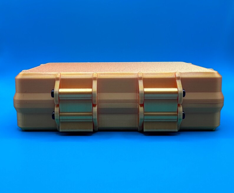 May include: A rectangular, gold-colored storage case with a textured top surface. The case features two prominent, hinged locking latches on the front side, each secured with small black pins, providing a secure closure for the container.
