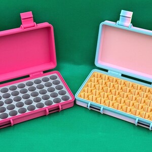 May include: Two rectangular storage cases with hinged lids. The left case is solid pink with a grey interior grid. The right case features a light blue frame, a pale pink interior lid, and a yellow interior grid with multiple circular slots.
