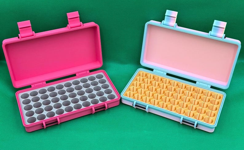 May include: Two rectangular storage cases with hinged lids. The left case is solid pink with a grey interior grid. The right case features a light blue frame, a pale pink interior lid, and a yellow interior grid with multiple circular slots.