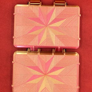May include: Two rectangular, hinged cases with a metallic gold finish and a pink, star-shaped geometric pattern on the lids. Each case features a textured surface with fine, radiating lines and two prominent gold-colored tabs at the top edge.