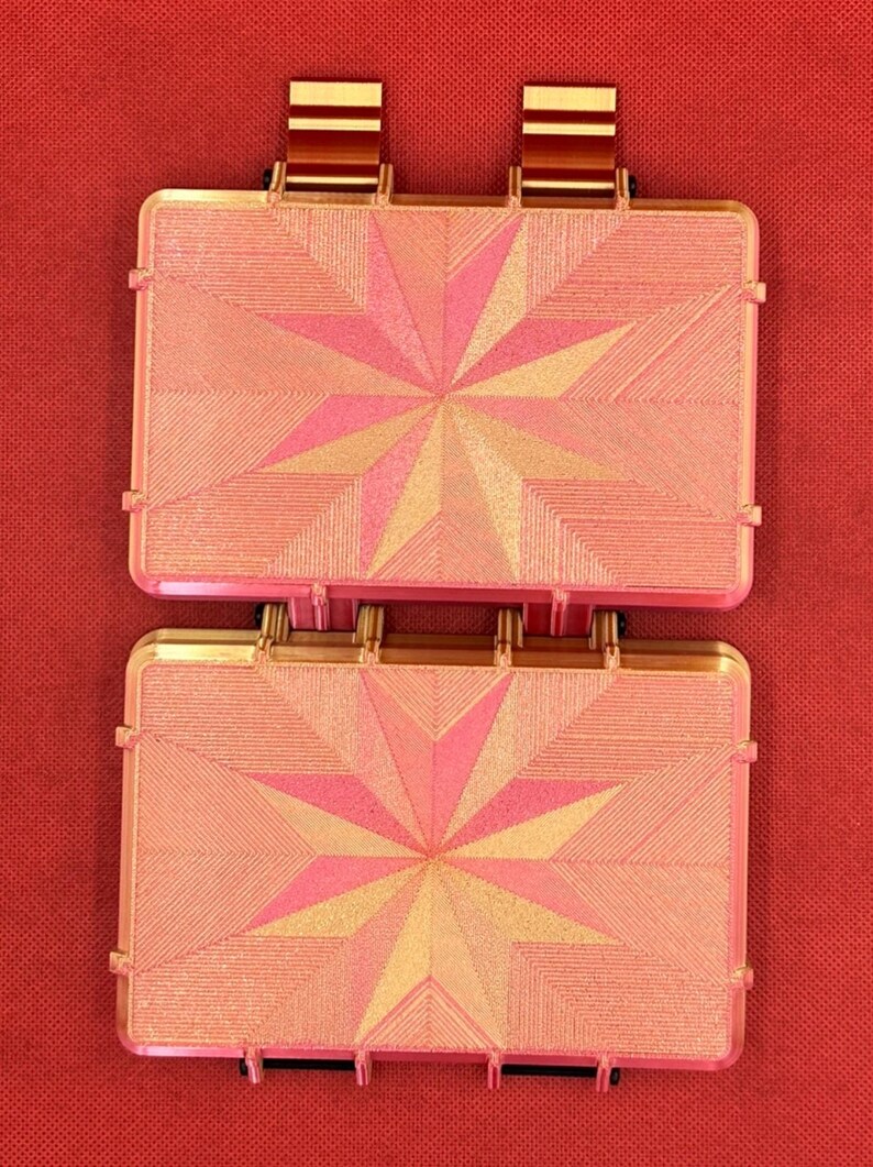 May include: Two rectangular, hinged cases with a metallic gold finish and a pink, star-shaped geometric pattern on the lids. Each case features a textured surface with fine, radiating lines and two prominent gold-colored tabs at the top edge.