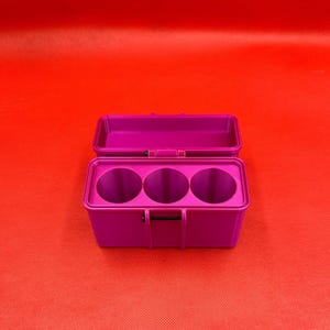 May include: A rectangular magenta storage case with a hinged lid in the open position. The interior of the base features three circular compartments arranged in a row, designed to hold cylindrical items securely within the container.