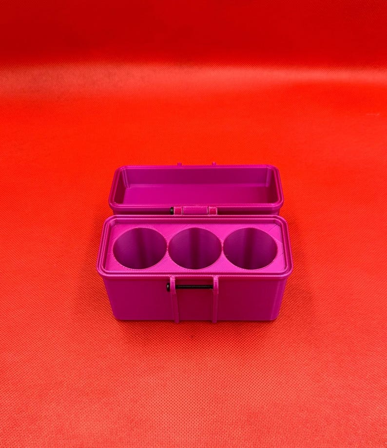 May include: A rectangular magenta storage case with a hinged lid in the open position. The interior of the base features three circular compartments arranged in a row, designed to hold cylindrical items securely within the container.