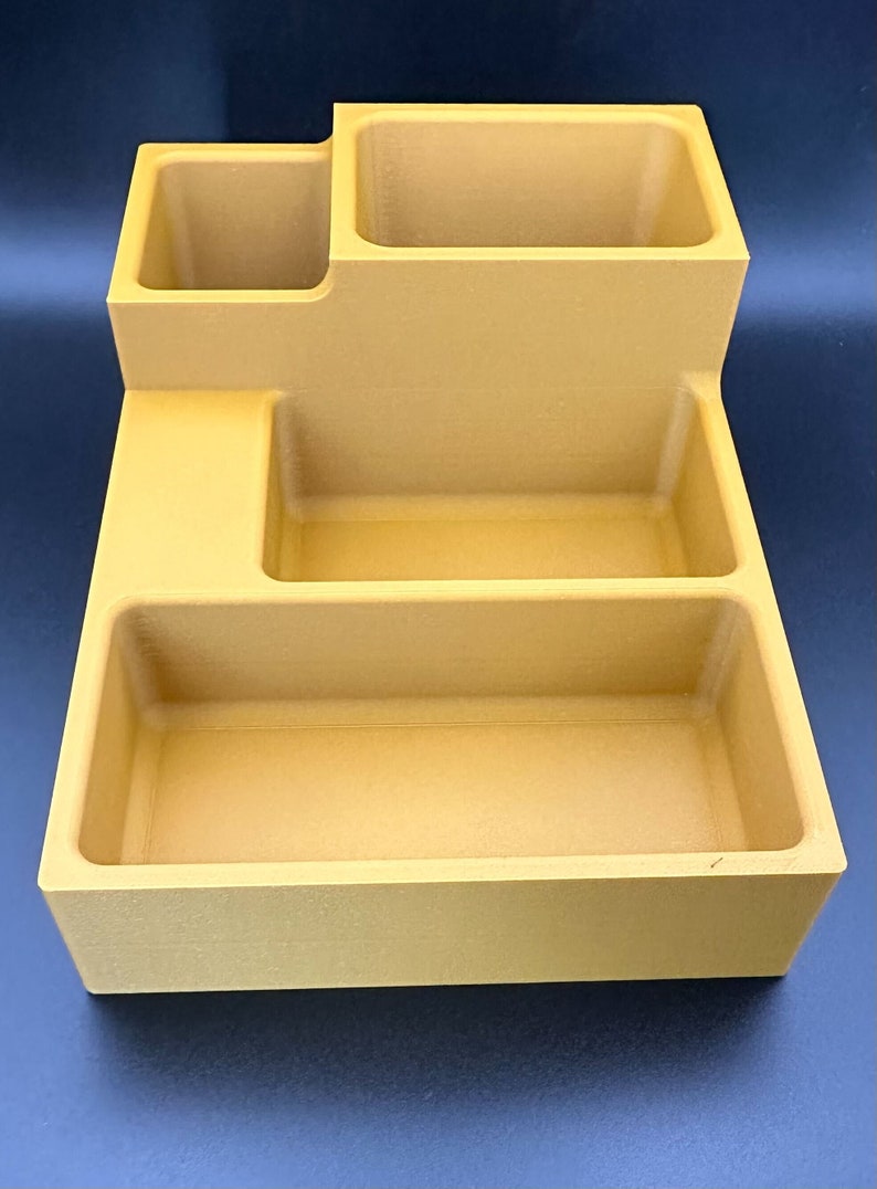 Op de afbeelding: Een gouden desktop organizer met vier vakken. De organizer heeft een rechthoekige vorm met een verhoogd gedeelte linksboven. De vakken zijn van verschillende groottes en diepten.