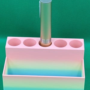 以下が含まれることがあります： ピンクとブルーのグラデーションのオーガナイザーで、ペンや鉛筆用のスロットが5つと、その他のアイテム用の大きなコンパートメントがあります。明るいブルーのトップが付いたゴールドのペンが、スロットの1つに入っています。