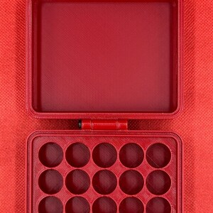 May include: A red rectangular storage case with a hinged lid and a gold-coloured metal latch. The interior of the base features a grid of twenty circular compartments arranged in four rows of five, while the lid interior is hollow.