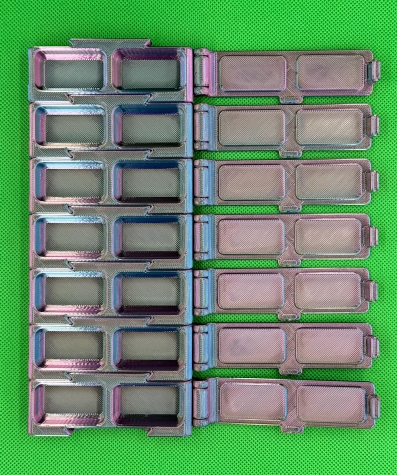 May include: A set of six iridescent, 3D-printed rectangular storage containers with hinged lids. Each container features two recessed compartments with a textured finish, displaying a color-shifting gradient of purple, blue, and metallic silver tones.