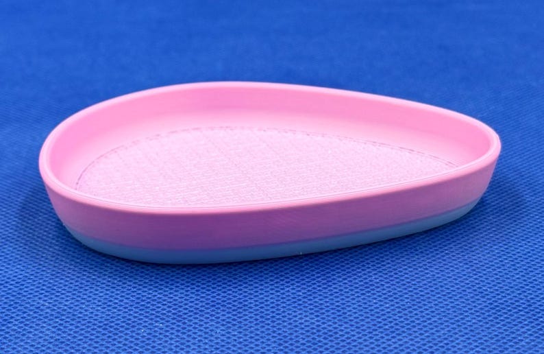 May include: A pink and blue silicone dish with a textured surface. The dish is shaped like a teardrop and has a raised edge.