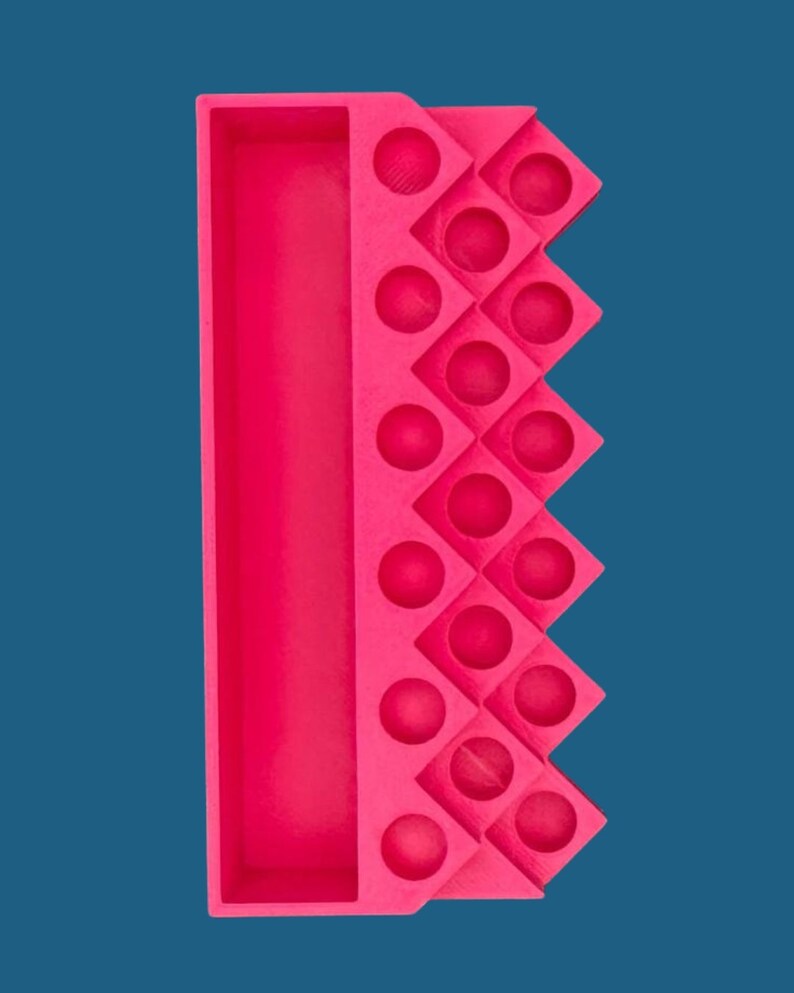 May include: A bright pink rectangular organizer tray featuring a long, deep storage compartment on the left side and a series of sixteen small, circular indentations arranged in a diamond-shaped grid pattern on the right side.