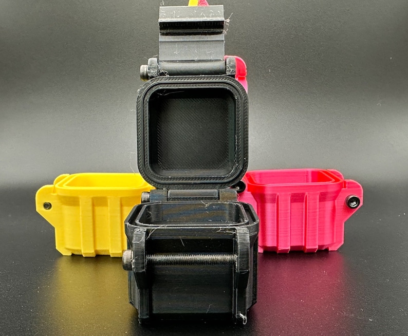 K&ouml;nnte beinhalten: Drei kleine, quadratische, 3D-gedruckte Aufbewahrungsbeh&auml;lter mit Klappdeckeln. Ein schwarzer Beh&auml;lter steht in der Mitte mit ge&ouml;ffnetem Deckel, w&auml;hrend ein gelber und ein pinker Beh&auml;lter jeweils dahinter auf beiden Seiten stehen.